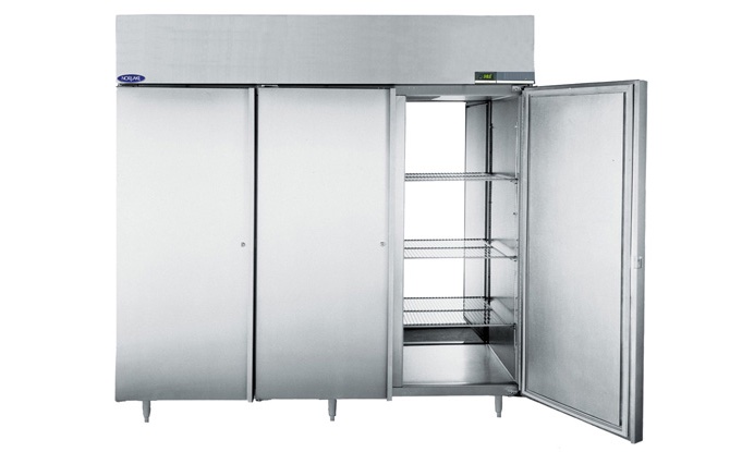 A large, three-door commercial stainless steel fridge stands on metal legs. One door is open, revealing multiple wire shelves inside, while the other two doors are closed. The appliance has a digital display panel on the upper right side.