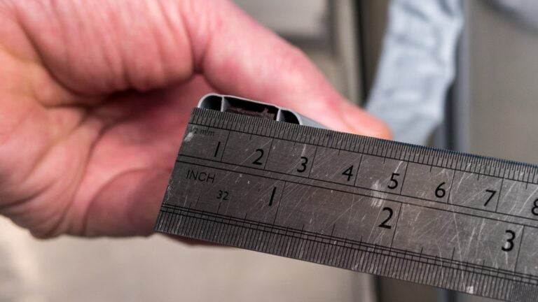 A close-up of a hand holding a metal ruler next to a plastic object, measuring its width. The ruler shows both inch and millimetre markings, with the plastic object’s width aligning to about 1.8 centimetres or 0.7 inches.