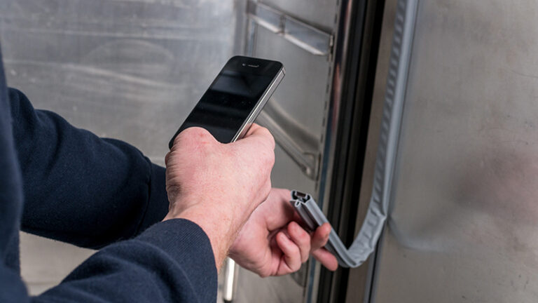A person holds a mobile phone in one hand and inspects or repairs a grey fridge or freezer door seal with the other hand. The person wears a dark long-sleeve shirt, and the scene appears to take place indoors near a metal surface.