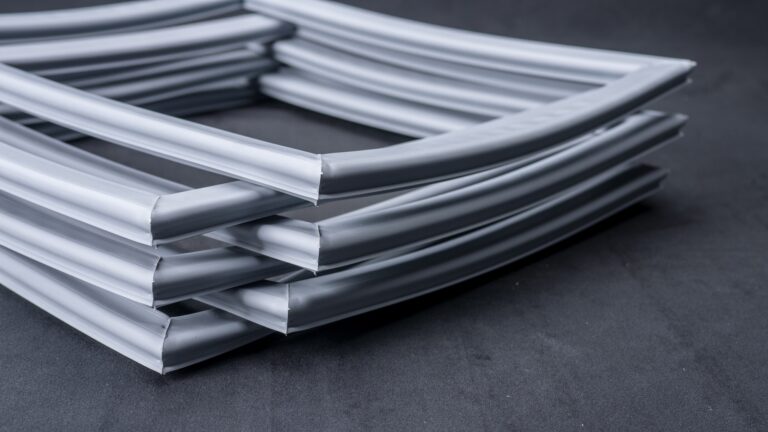 A close-up of several stacked, curved, silver-grey rubber or plastic strips with hollow centres, arranged neatly on a dark, textured surface. The material is smooth and flexible, and the strips form a geometric pattern.