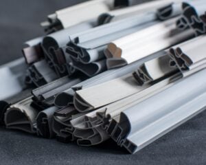 A close-up of a pile of various grey and black rubber or plastic draught excluder strips, showing different cross-sectional profiles and textures, placed on a dark surface. The materials are stacked in an overlapping, disordered arrangement.