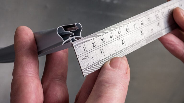 A close-up of hands holding a metal ruler against the cross-section of a grey rubber or plastic strip, measuring its width in centimetres and inches. The focus is on the measurement, with the numbers clearly visible on the ruler.