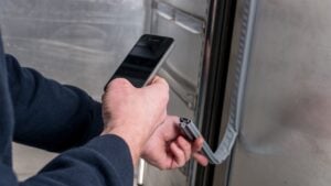 A person holds a mobile phone in one hand and inspects a damaged rubber seal on the edge of a stainless steel door or appliance with the other hand, possibly documenting the damage for repair or maintenance.