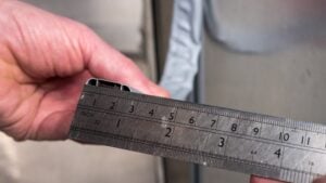 A close-up of a hand holding a metal ruler marked in inches and centimetres, positioned against a metallic surface, with a wavy line of white sealant visible in the background. The ruler’s measurements are clearly visible.
