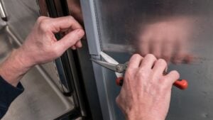 A person uses red-handled scissors to trim a grey rubber gasket on the edge of a metal fridge door. One hand holds the gasket while the other cuts, with the reflective stainless steel surface of the fridge visible in the background.