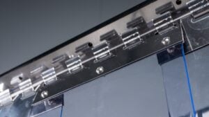 Close-up of a metallic hinge mechanism attached to a transparent, blue-tinted plastic sheet. Several screws and metal tabs are visible, reflecting light. The background is out of focus, emphasising the structure and materials of the hinge assembly.