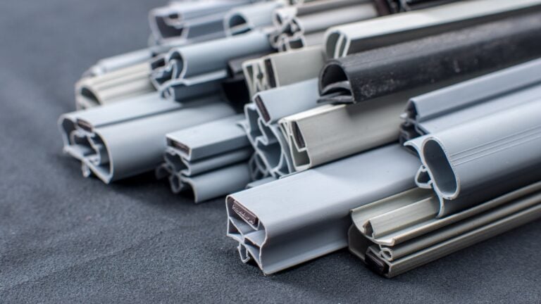 Close-up of stacked grey and black rubber or plastic weatherstrips with varied profiles and cross-sections, displayed on a dark textured surface. The strips have hollow and solid portions, and are arranged in an overlapping, clustered manner.