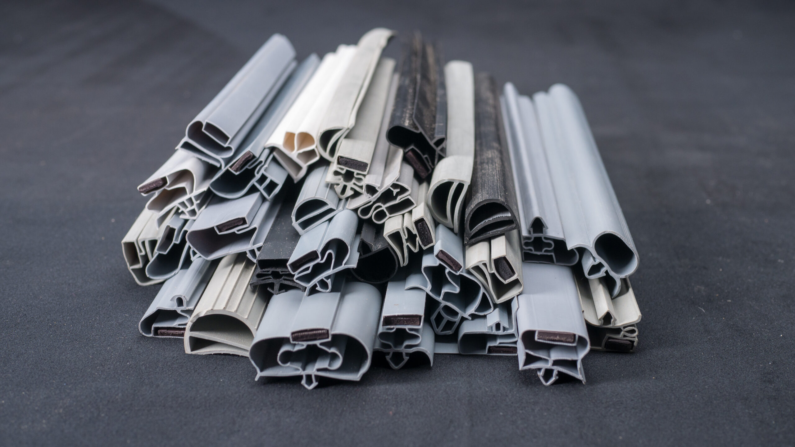 A pile of various grey and black rubber or plastic weatherstripping seals, neatly arranged on a dark surface, showing different shapes and cross-sections, likely used for doors or windows to prevent draughts and water leakage.