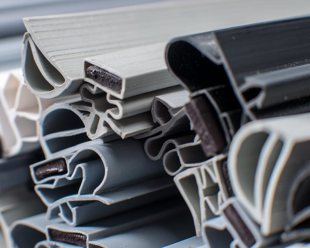 Close-up of stacked, variously shaped grey and black rubber and plastic weatherstripping seals, showing intricate cross-sections and textures. The materials are layered and interlocked, revealing their flexible and functional design for insulation.