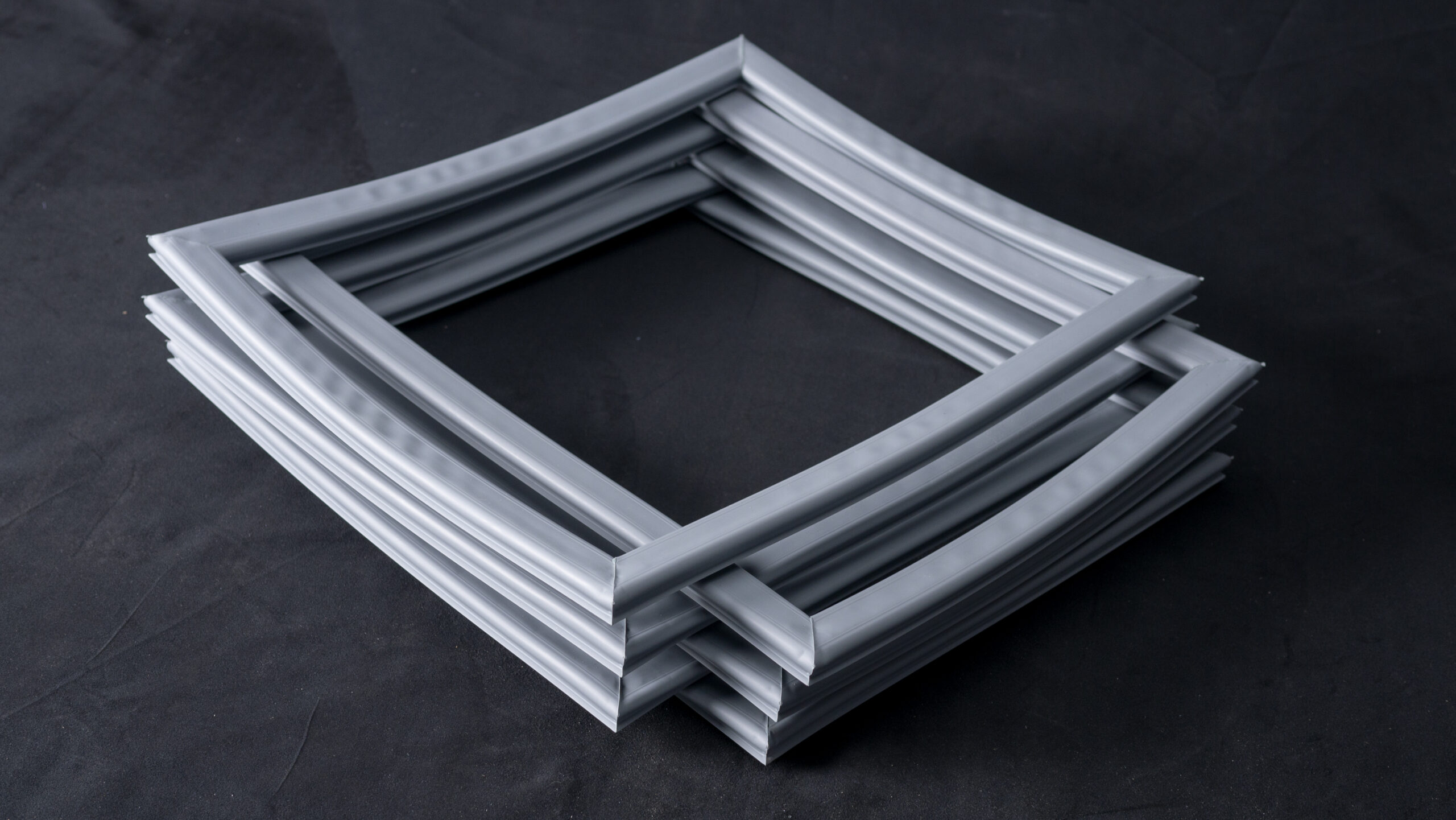 Four grey rectangular frames are stacked diagonally on a dark surface, creating a twisted, layered look. The frames appear to be made of soft, flexible material, with a smooth texture and slightly wavy edges.