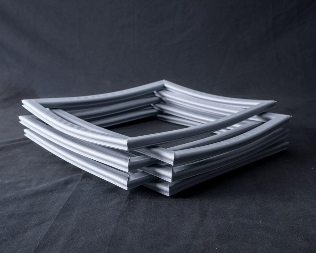 A stack of four square, grey, flexible rubber gaskets arranged neatly on a dark fabric background. The gaskets have a groove along their length and are stacked in an alternating, offset pattern.