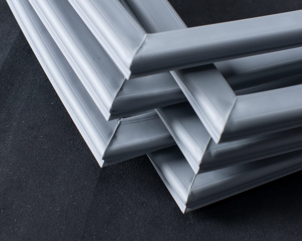 Three grey picture frames intersect and overlap at varying angles on a black surface, forming a geometric arrangement with their corners. The frames create a layered, abstract pattern with smooth, matte finishes and clean lines.