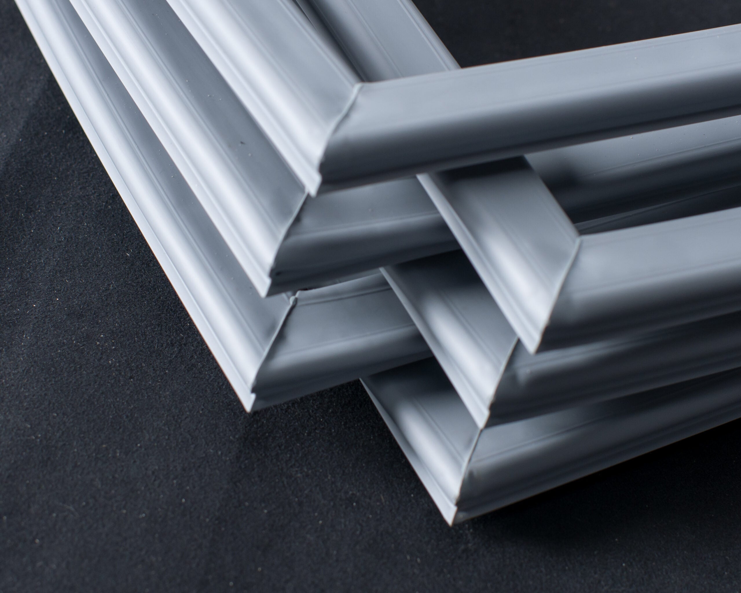Three grey picture frames intersect and overlap at varying angles on a black surface, forming a geometric arrangement with their corners. The frames create a layered, abstract pattern with smooth, matte finishes and clean lines.