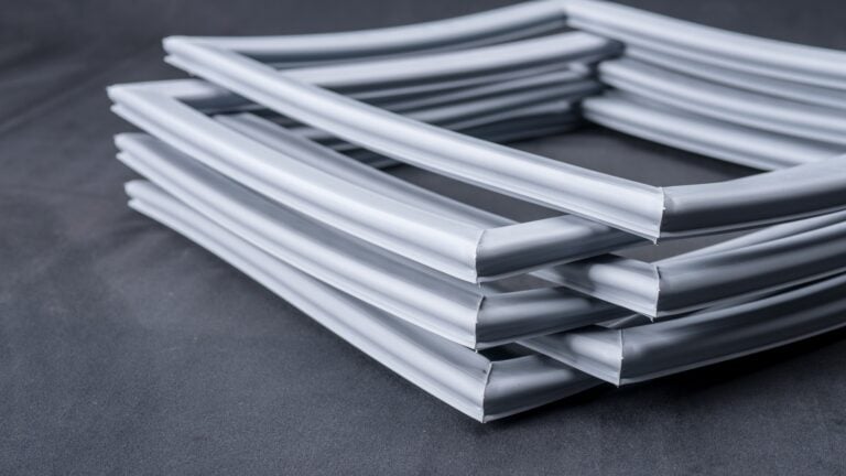 Four rectangular, grey rubber or plastic gaskets are stacked in an alternating criss-cross pattern on a dark grey surface. The gaskets have smooth surfaces and rounded edges, forming an orderly, geometric arrangement.