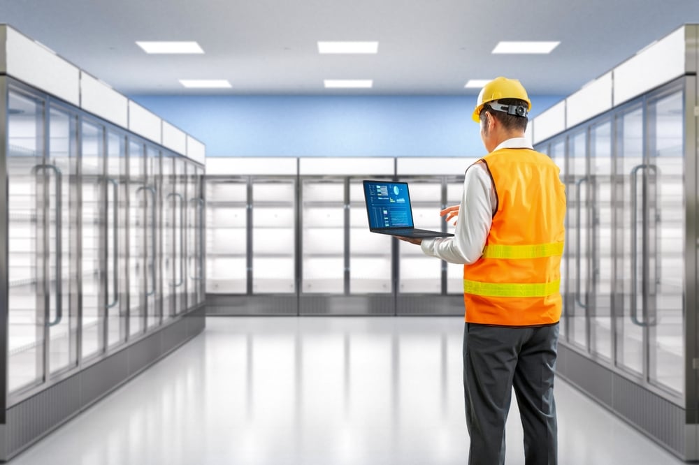A man in a yellow hard hat and orange safety vest stands in a brightly lit, empty supermarket aisle with glass-door fridges, holding and working on an open laptop, possibly inspecting or managing stock or equipment.