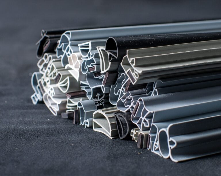 Several aluminium extrusions with complex cross-sectional shapes are stacked on a dark surface. The metal pieces are in various shades of grey, black, and silver, and are organised in a slightly messy, overlapping arrangement.