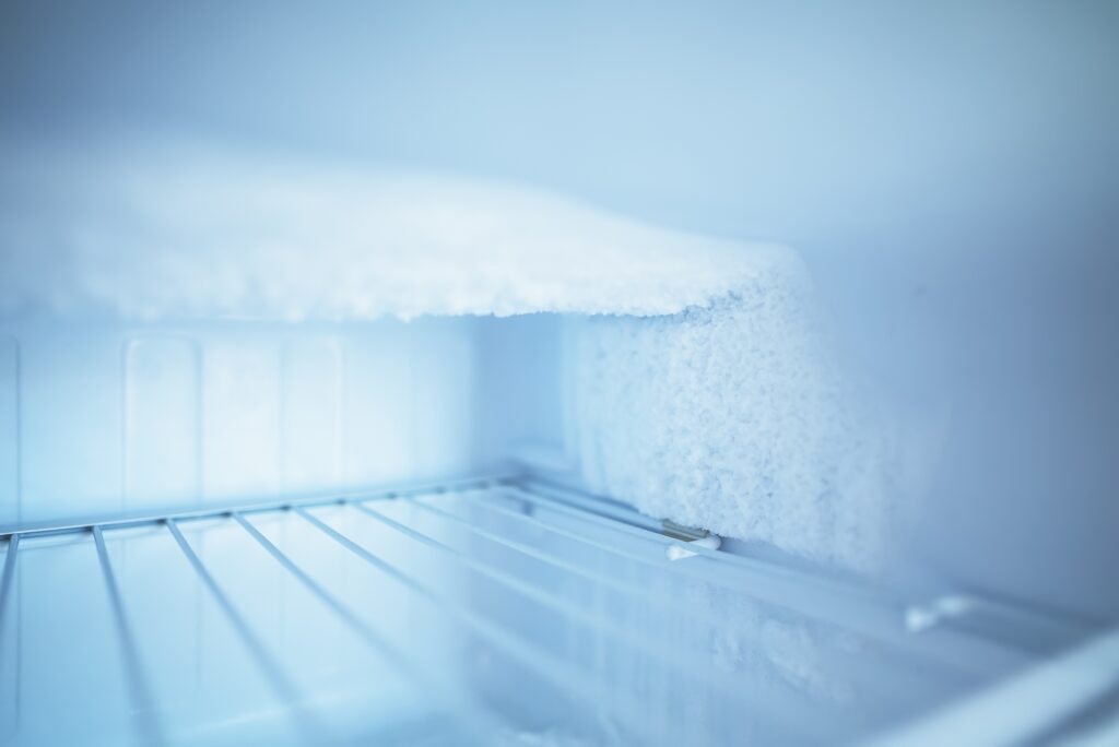The inside of a freezer with a thick layer of white frost and ice build-up on the top and back wall, above empty metal shelves. The scene is lit with a cool, bluish light, emphasising the cold, icy environment.