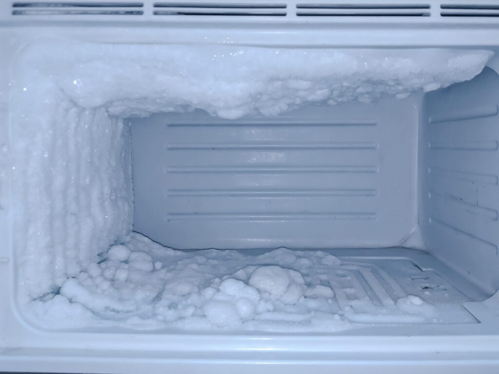 An empty freezer with heavy frost and ice build-up on the back, left wall, and floor. The thick layer of white ice appears uneven and jagged, coating much of the interior surfaces, indicating the freezer needs defrosting.
