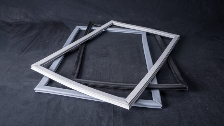 Three empty rectangular picture frames, two white and one black, are stacked unevenly on a dark, textured surface with a dark backdrop. The frames overlap each other, creating a layered effect with visible shadows and highlights.