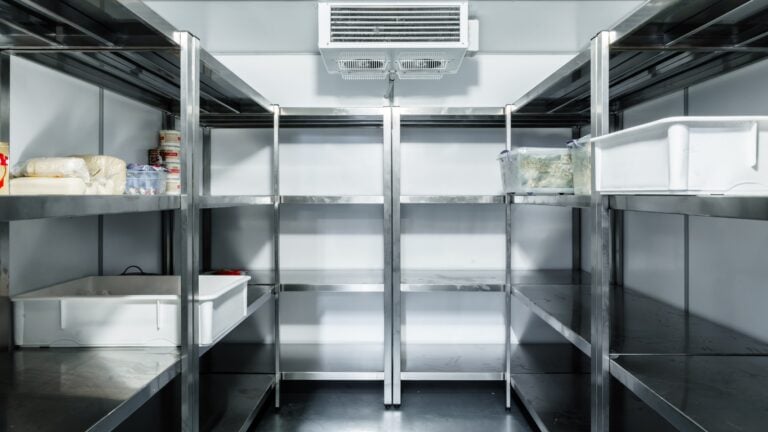 A clean, empty commercial walk-in fridge with stainless steel shelves along both sides. A few plastic bins and containers with food items are sparsely placed on the shelves, while an air vent helps minimise cold room condensation on the back wall.