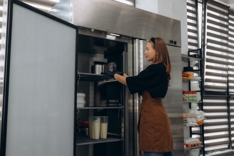 Woman baker arranging containers and ingredients inside a commercial stainless steel refrigerator in a modern kitchen