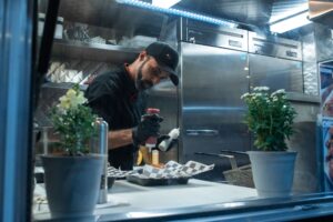 Man dispensing ketchup onto hot dog in commercial kitchen, industrial fridges visible in background